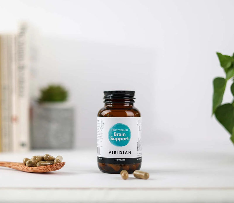 Bottle of viridian brain support multivitamin capsules on a white surface with a plant and a wooden spoon with cloose capsules