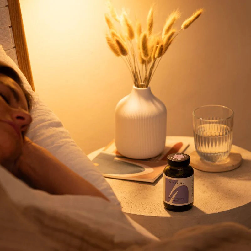 Person lying in bed with a nightstand featuring a vase, glass, and bottle.