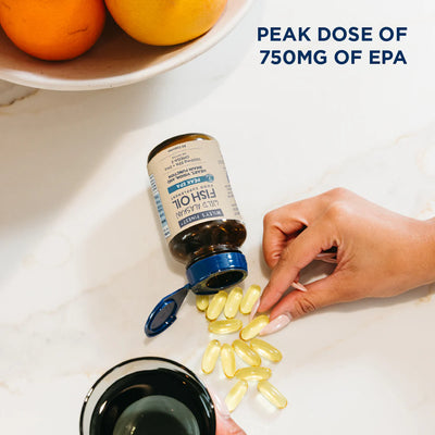 Hand pouring fish oil capsules from a bottle onto a white surface with oranges in the background.