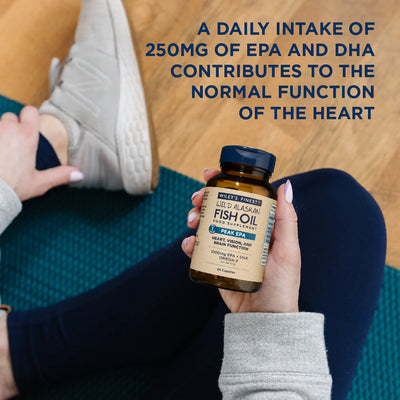 Person holding a bottle of Wiley's Finest Wild Alaskan Fish Oil supplement on a wooden floor.