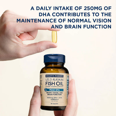 Hand holding a bottle of Wiley's Finest Wild Alaskan Fish Oil with a pill above it, on a light background.