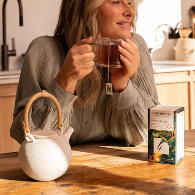 Woman holding a mug of tea with a teapot and a box of tea on a kitchen counter.