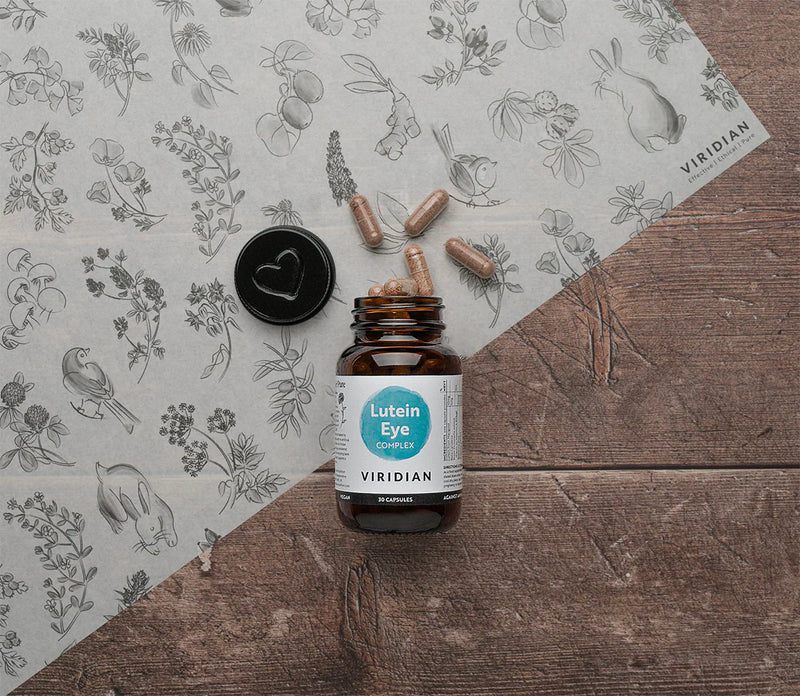 Bottle of Viridian Lutein Eye Complex with capsules on a wooden surface and patterned paper background.