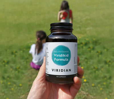 Hand holding a bottle of Viridian Multivitamin Viridikid Formula with children playing in the background.