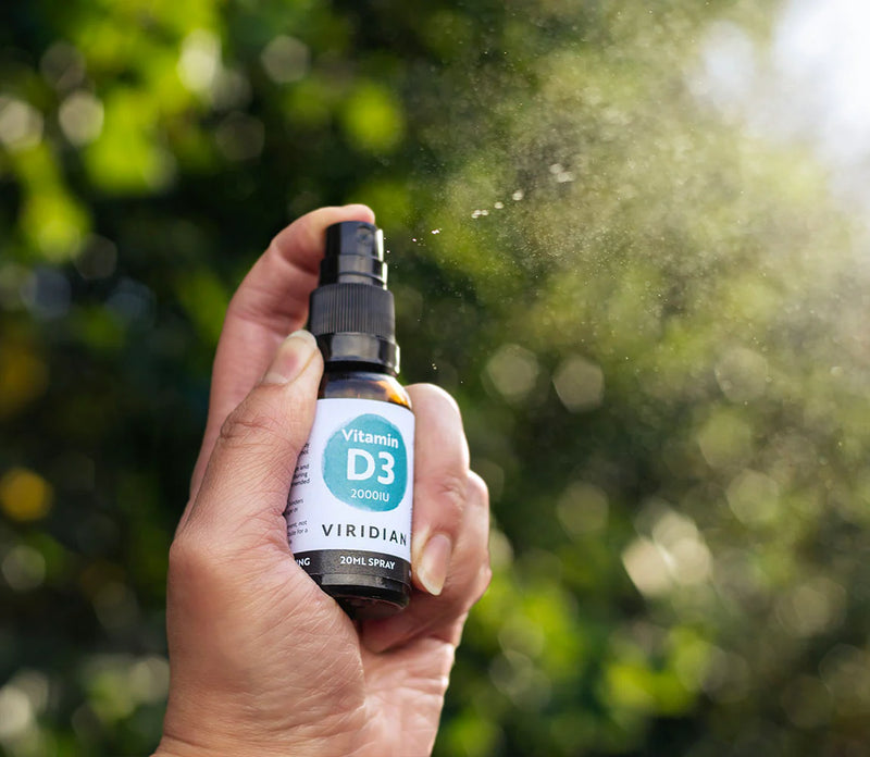 Hand holding a Viridian Vitamin D3 spray bottle with a blurred green outdoor background
