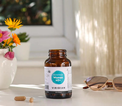 Bottle of viridian woman 40+ multivitamins  on a white surface with loose capsules , sunglasses and a vase of flowers